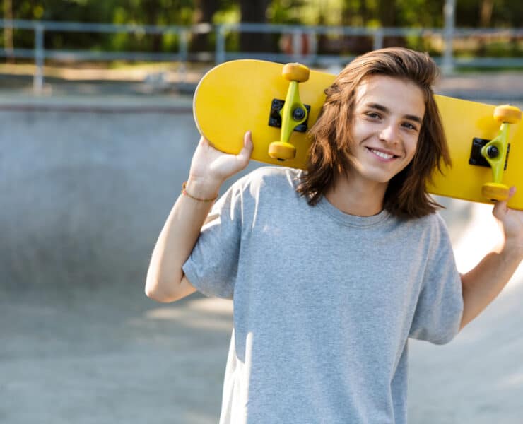 a teenager holding a yellow skateboard behind his head, smiling