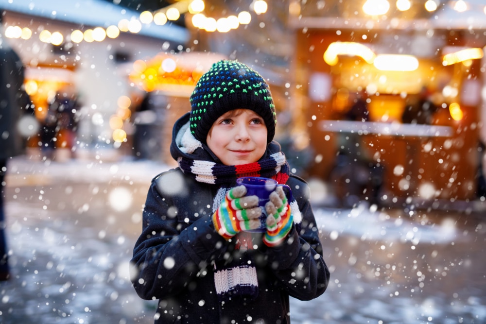 little boy holding cup of hot chocolate in the snow at a christmas market