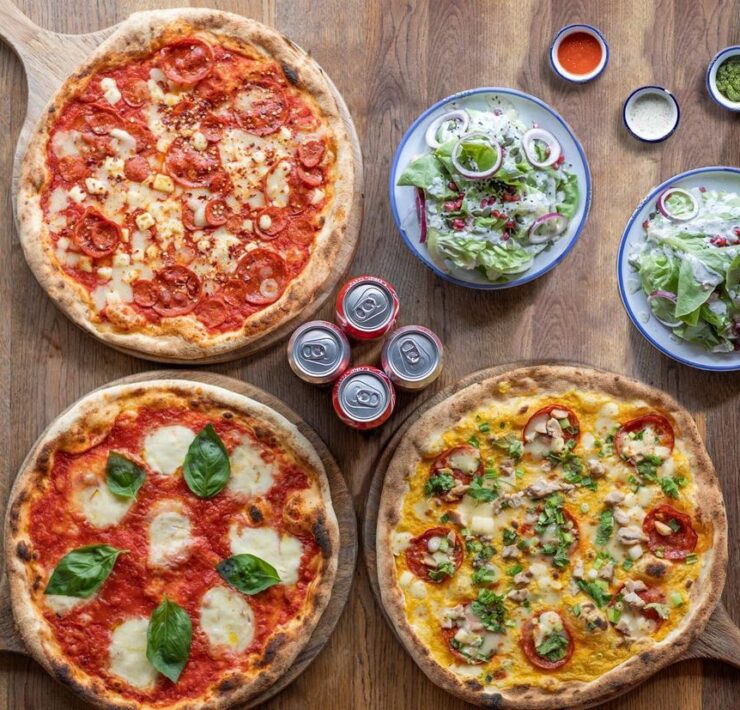 three pizzas on a wooden plate with drinks and side salads next to it.