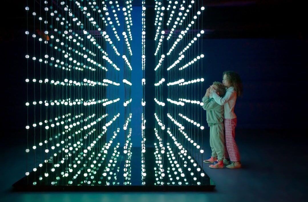 girl covering boys eyes next to a light installation at twist museum london