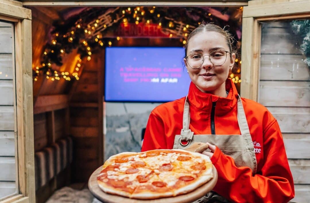 bar hutte worker holding out a large pizza in front of alpine hut