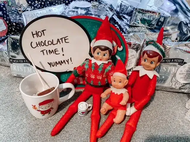 elf on the shelf elves sitting next to a mug with hot chocolate in it with a speech bubble sign that says hot chocolate time!