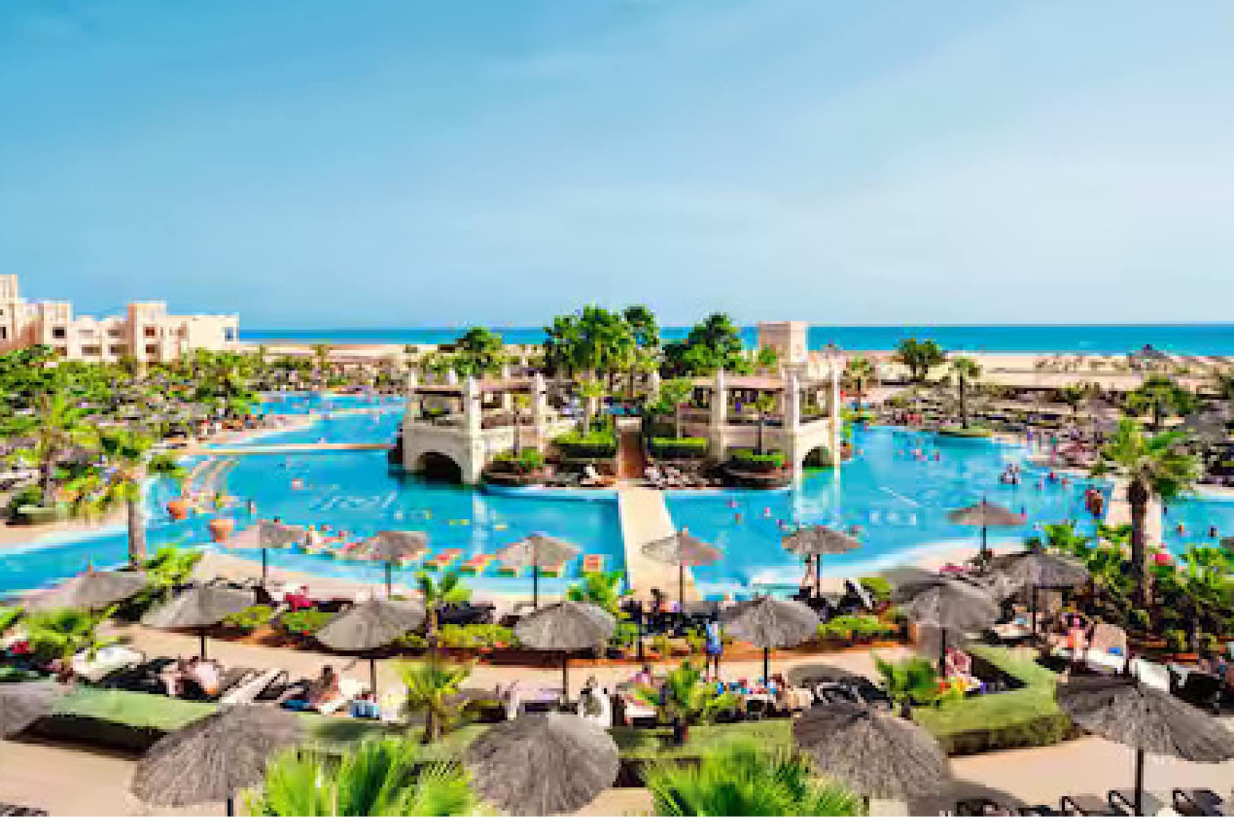 hotel with pool surrounding it and green trees with the blue ocean in the background