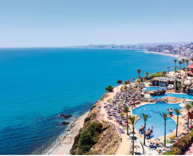 birds eye view of ocean next to a resort with palm trees and a pool