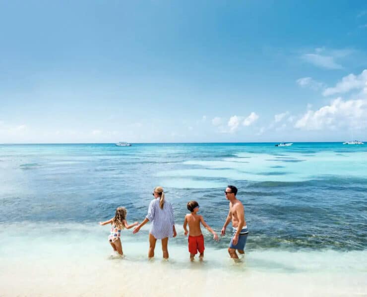 A family of four stands in the shallow, turquoise waters of a sunny beach in the Dominican Republic