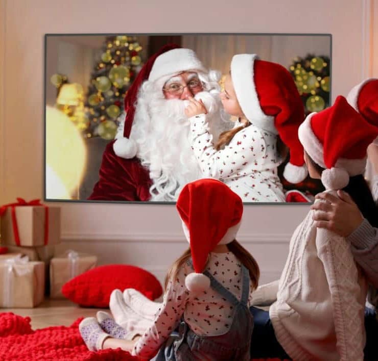 A family wearing festive red hats sits on the floor watching a programme featuring Father Christmas in a decorated lounge