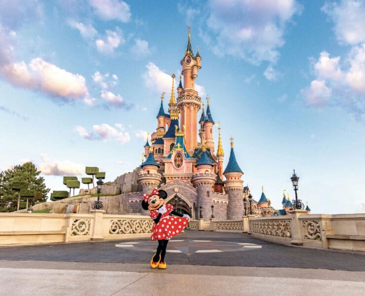Minnie Mouse stands in her classic red polka dot dress on the stone bridge in front of Sleeping Beauty Castle at Disneyland