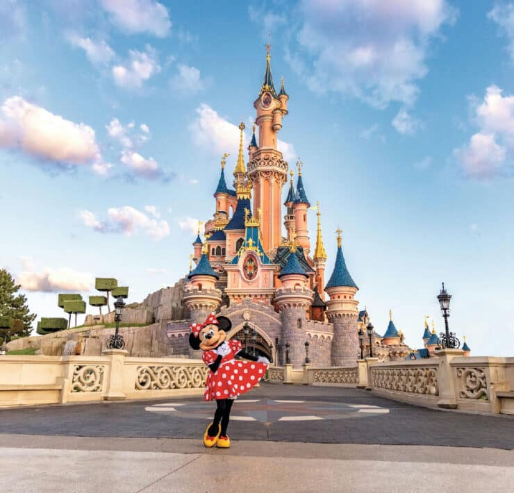 Minnie Mouse stands in her classic red polka dot dress on the stone bridge in front of Sleeping Beauty Castle at Disneyland