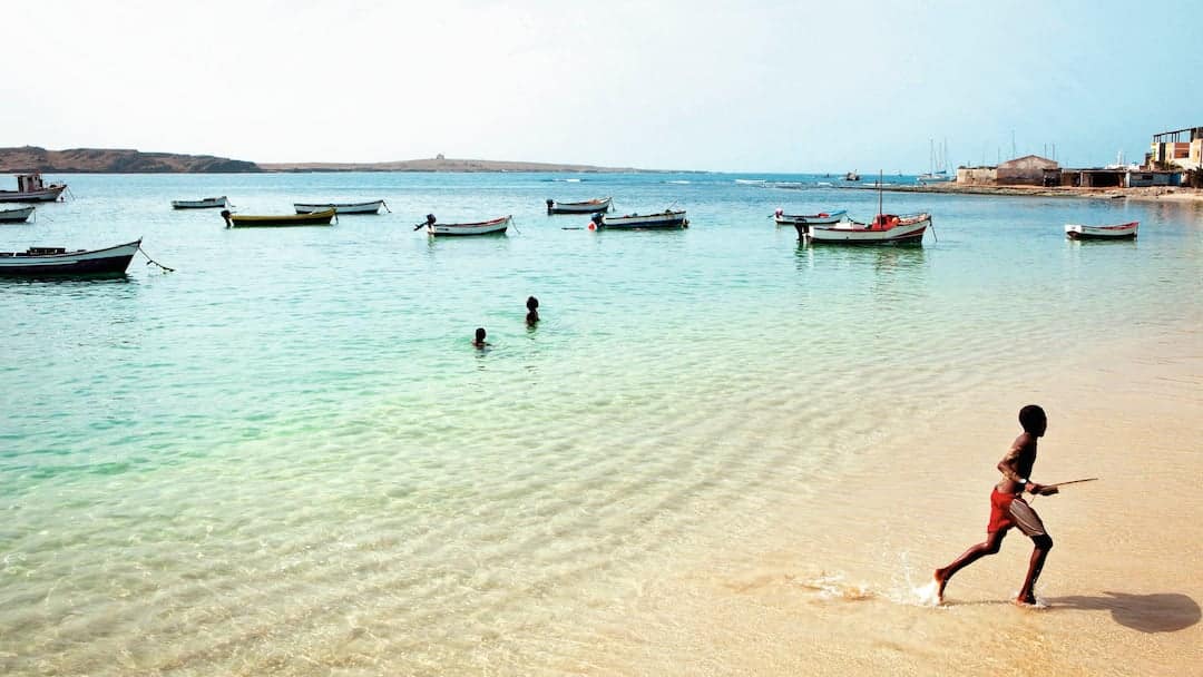 A boy in red shorts runs along a sandy beach towards clear turquoise water where several small fishing boats are moored