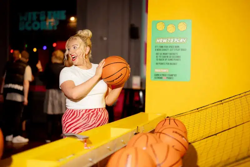 A cheerful young woman with blonde hair in a bun holds a basketball at an indoor arcade game