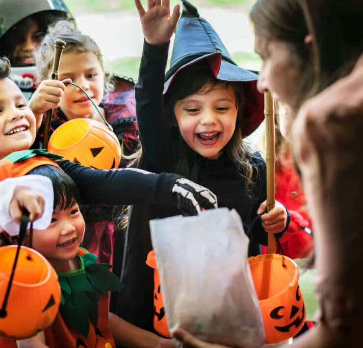 A group of young children in fancy dress, including a witch and a skeleton, enjoy trick-or-treating at a doorstep during